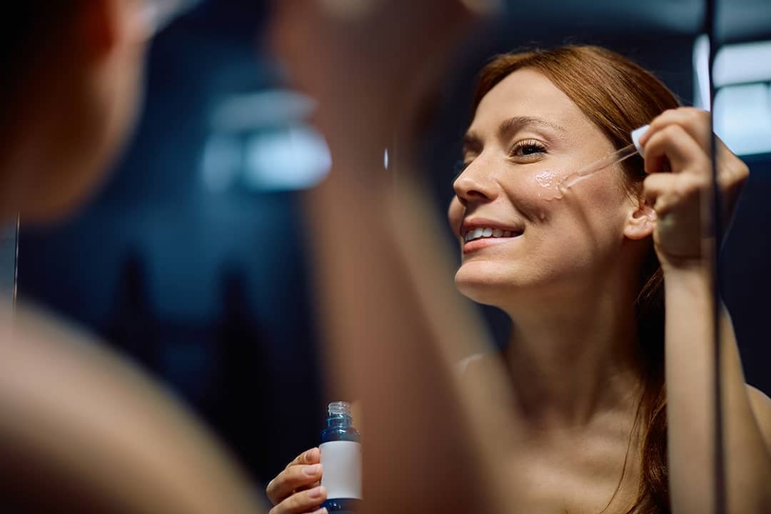 A person applying a moisturizing facial serum with a dropper in front of a mirror.